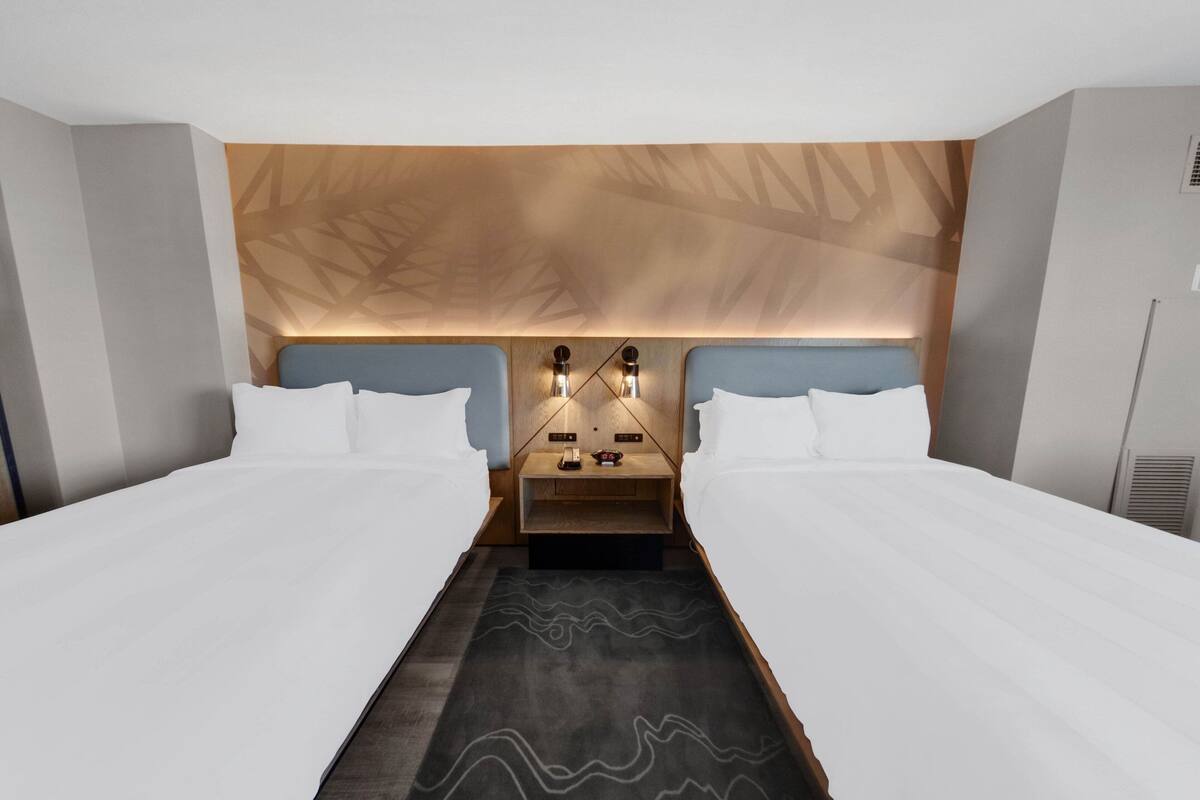 A modern hotel room with two beds, a wooden headboard, a nightstand with a lamp, and a patterned rug.