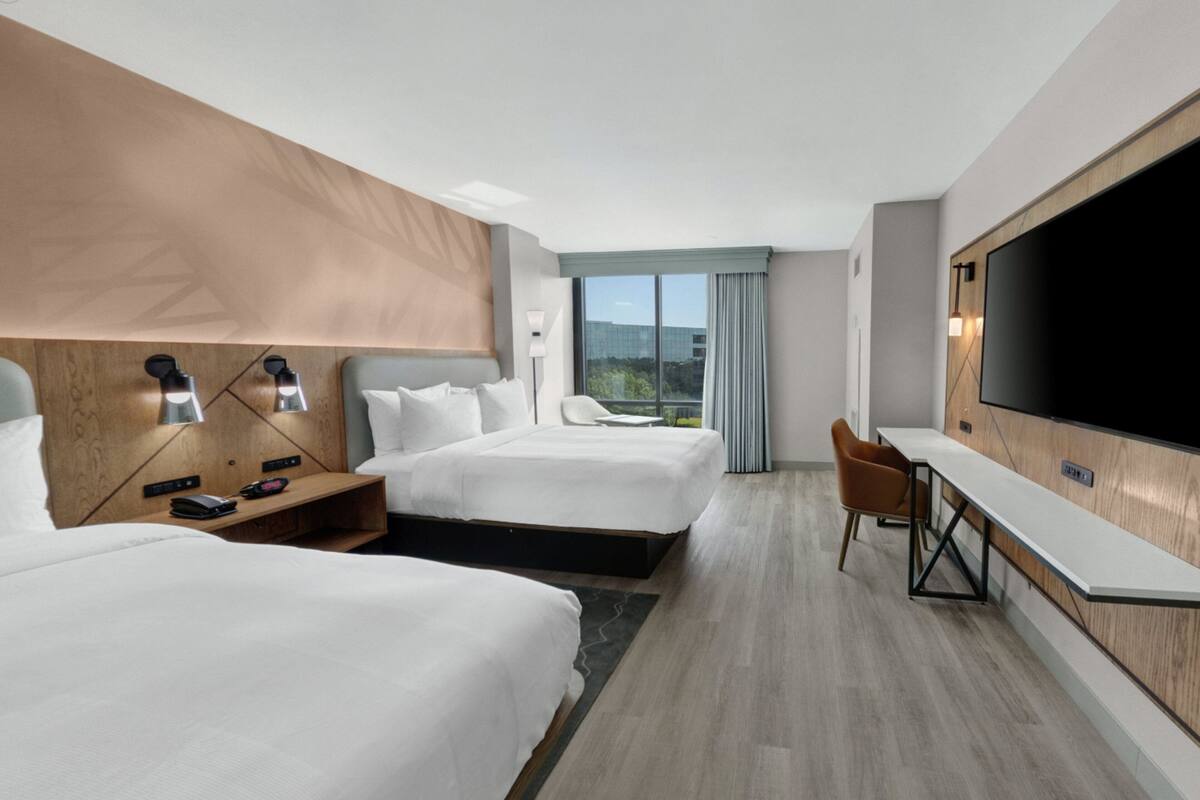 A modern hotel room with two beds, a desk, a TV, and a view of the outdoors.