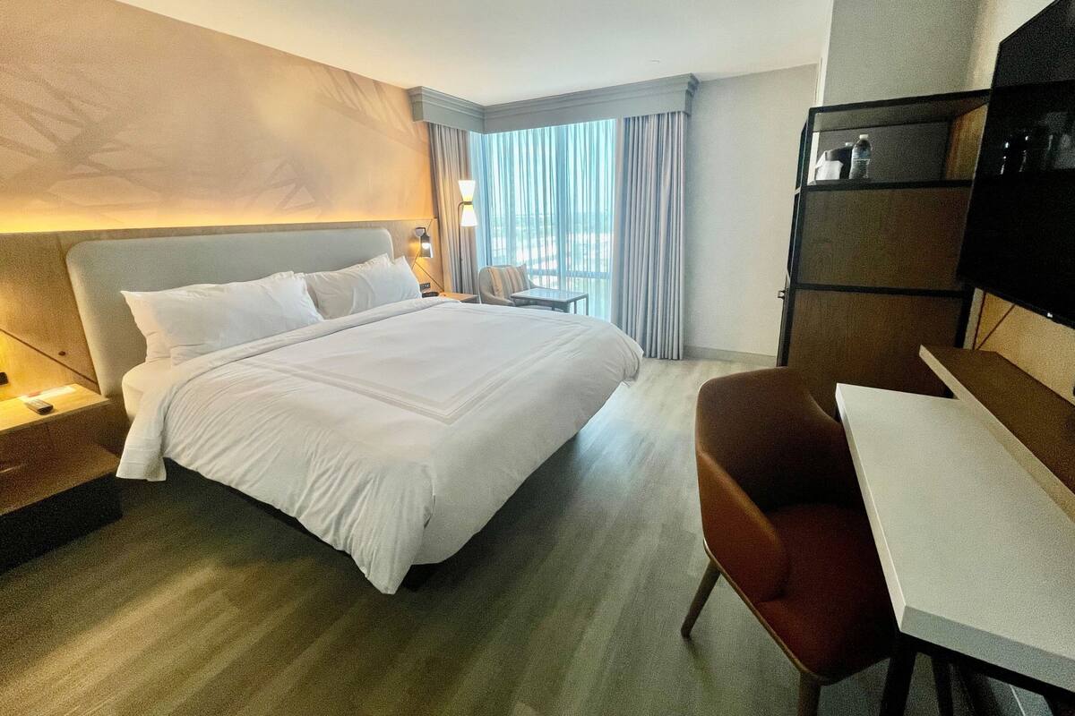 A hotel room with a large bed, a desk, a chair, a TV, and a view of the outdoors through the window.