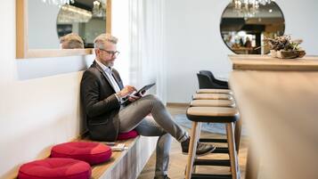 Een man zittend op een bankje in een modern interieur, met houten banken, rode kussens en een spiegel die een kroonluchter weerspiegelt.