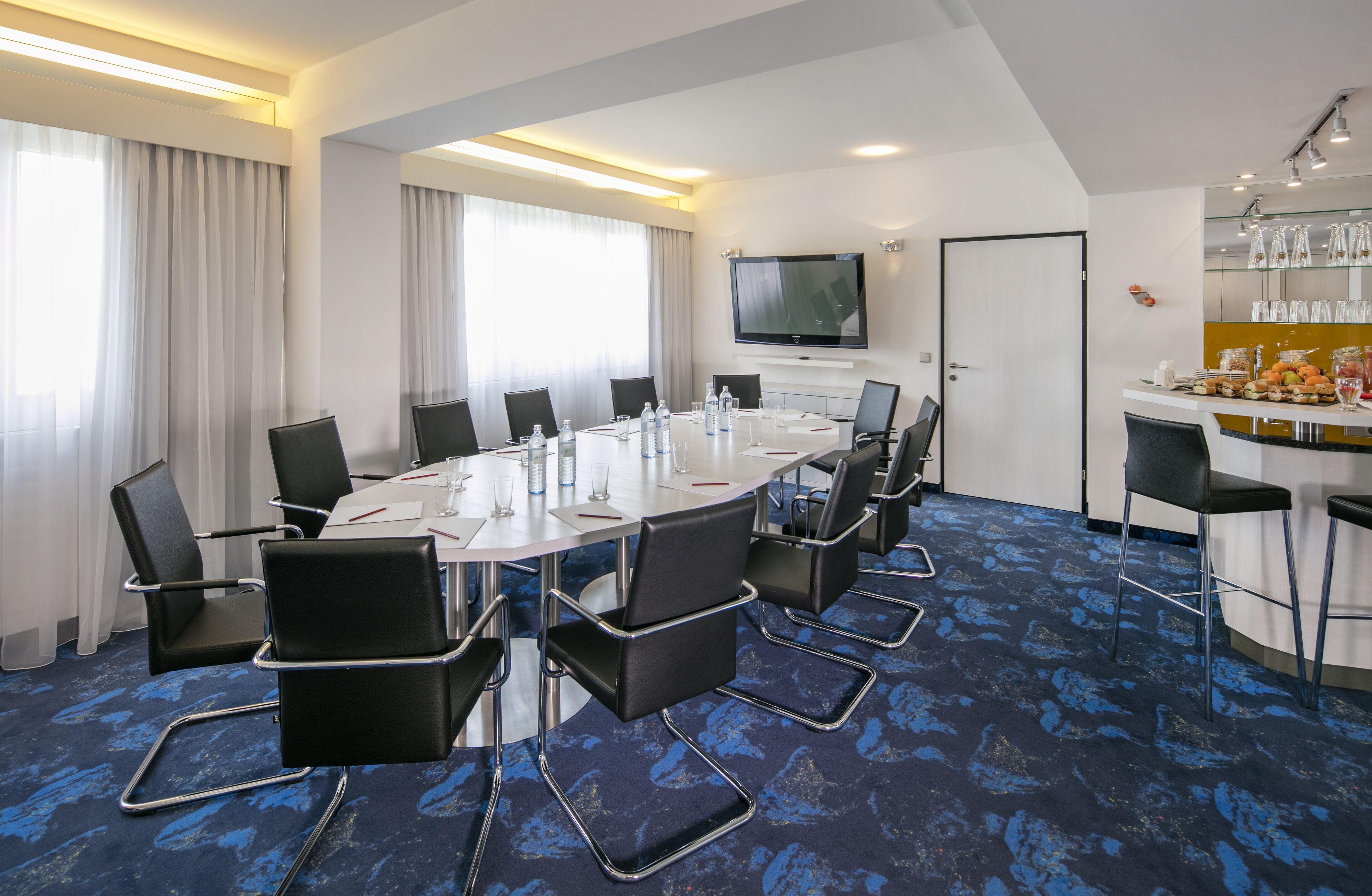 Un salón de conferencias con una mesa redonda, sillas negras, un televisor de pantalla plana y una zona de buffet con comida y bebidas.