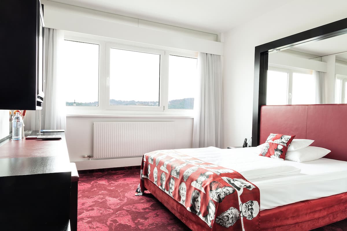 A hotel room with a large bed, a desk, a TV, and a window with a city view.