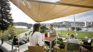 An outdoor seating area with tables and chairs, overlooking a body of water and a cityscape. - ARCOTEL Nike (Linz)