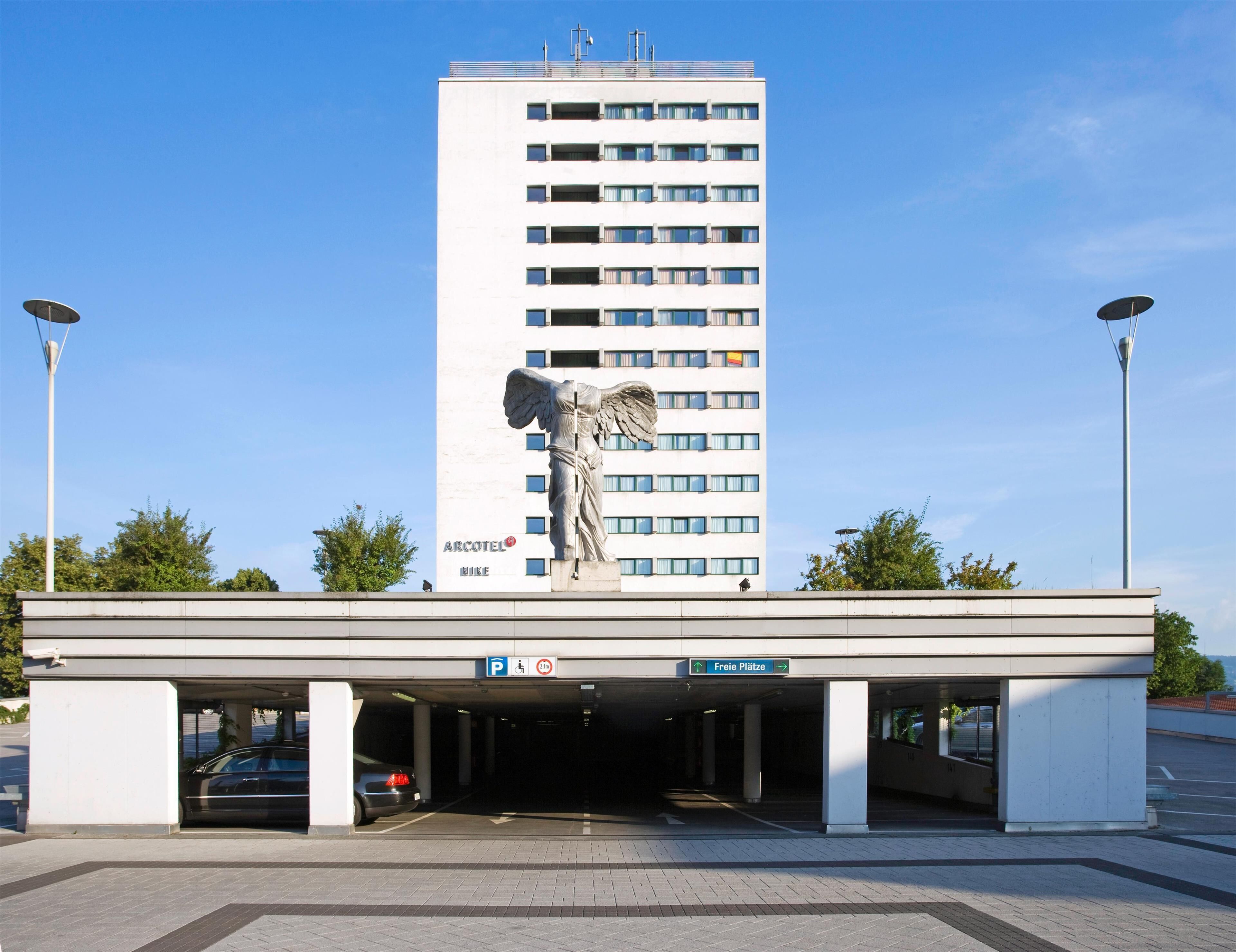 Einfahrt einer Parkgarage mit einer Statue eines Engels, einem Schild mit der Aufschrift 'ARCOTEL NIKE' und einem Parkplatzschild mit einem 'P'.
