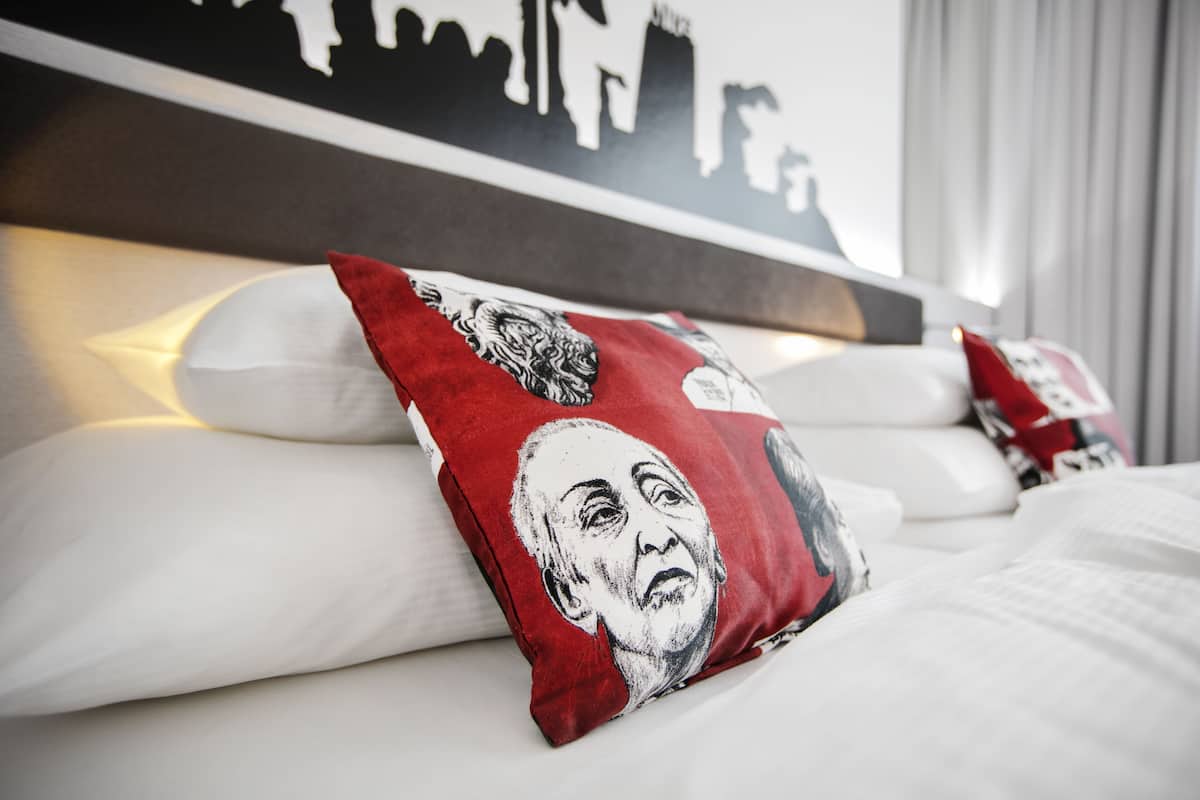 A hotel room with a neatly made bed, pillows with a red and white design, and a city skyline silhouette on the headboard.