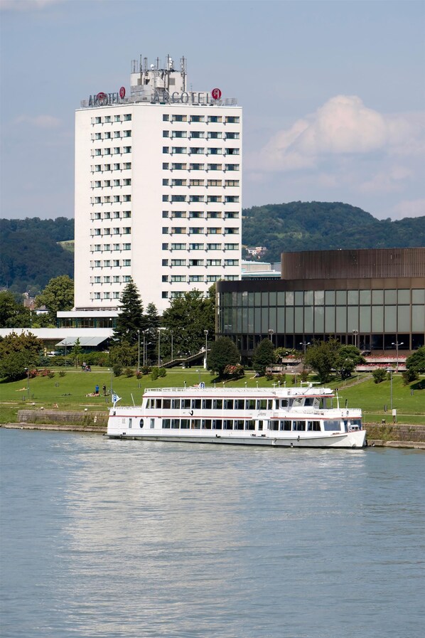 A large white hotel with multiple floors and balconies, a modern building with a flat roof, and a river with a white boat.