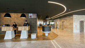 A modern reception area with wooden reception desks, a central marble sculpture, and a herringbone floor pattern. - ARCOTEL Nike (Linz)