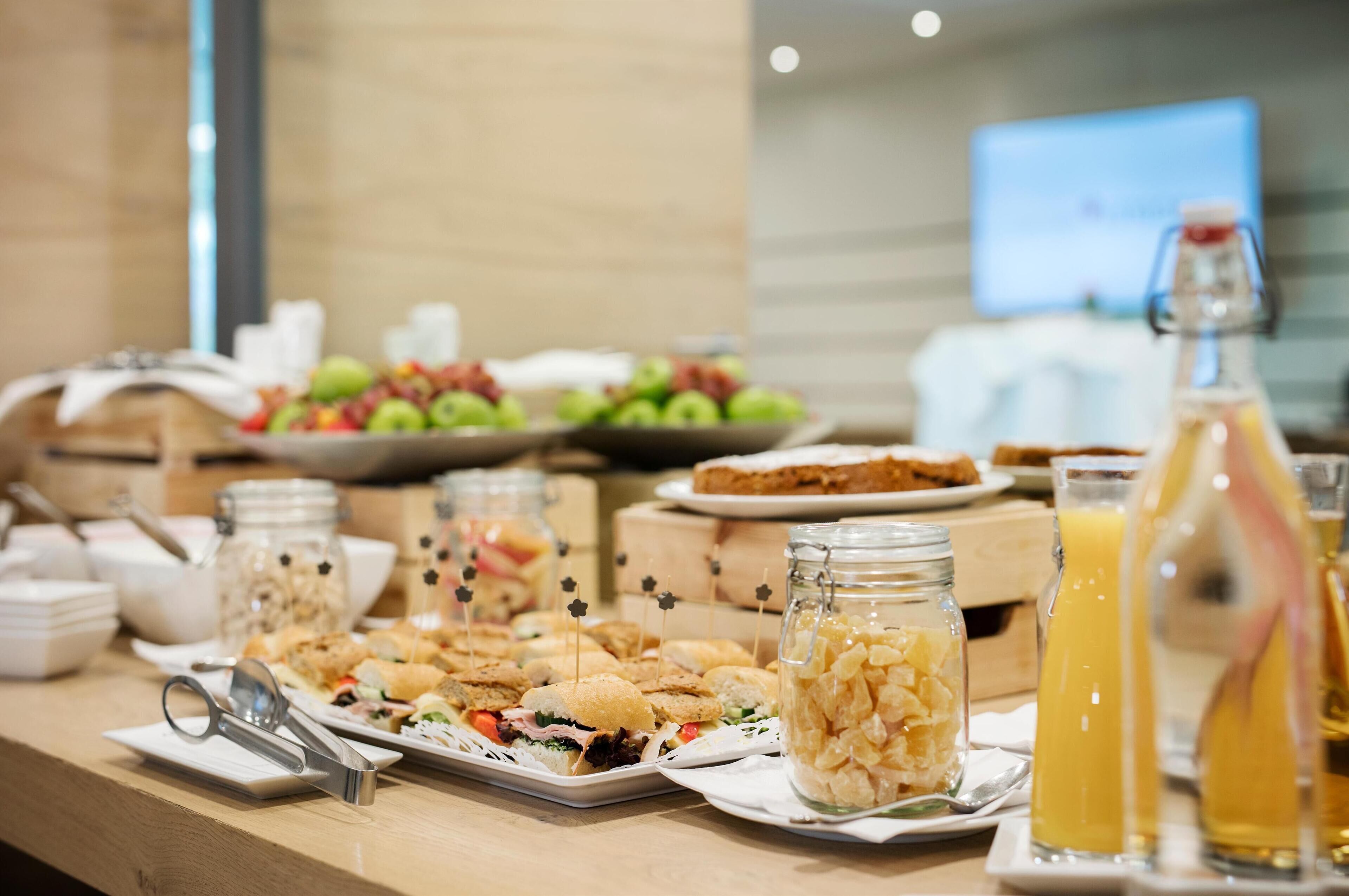 Ein Buffet mit verschiedenen Speisen, darunter Sandwiches, Salate und Obst. Es sind außerdem mehrere Glasbehälter und Flaschen zu sehen, vermutlich mit Soßen oder Getränken.