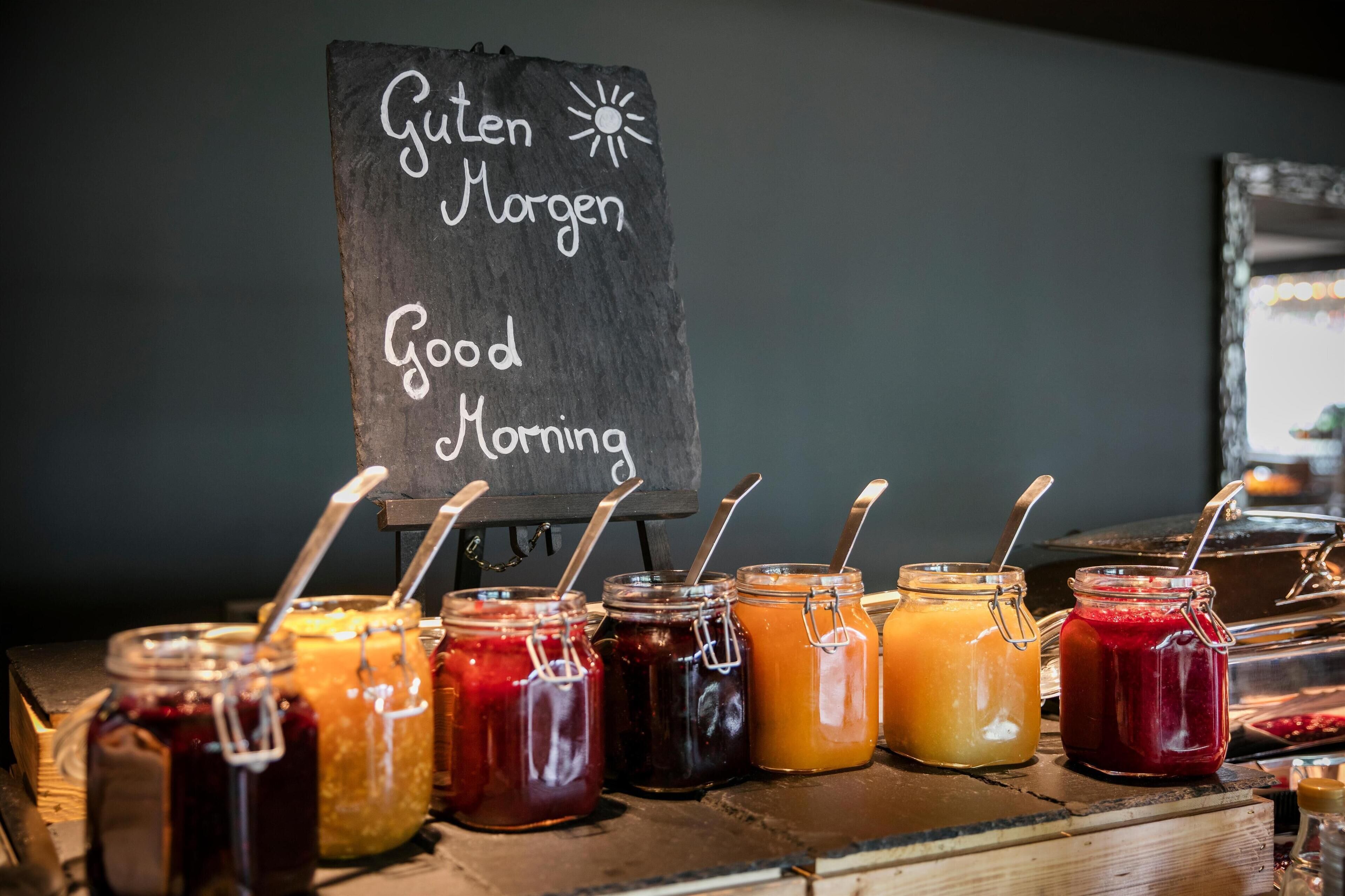 Una barra de desayuno con un letrero de pizarra que dice 'Guten Morgen Good Morning' y varios frascos de mermeladas y conservas.