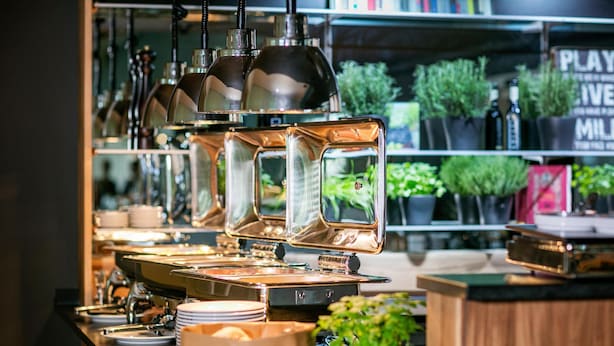 A buffet setup with multiple serving stations, a variety of dishes, and a background with shelves containing plants and a sign. - ARCOTEL Nike (Linz)