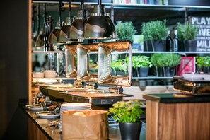 A buffet setup with multiple serving stations, a variety of dishes, and a background with shelves containing plants and a sign.