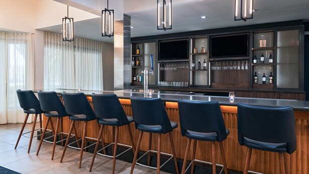 A modern bar area with a long counter, multiple bar stools, and a television screen. - Courtyard by Marriott San Antonio Airport (San Antonio)