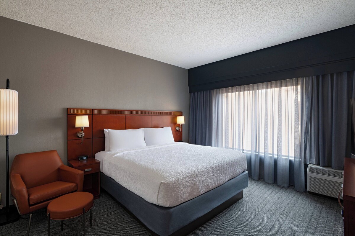 Ein Hotelzimmer mit einem großen Bett, einem Holzkopfstück, einem Nachttisch mit Lampe, einem Ledersessel und Blick ins Freie durch ein Fenster mit Vorhängen.