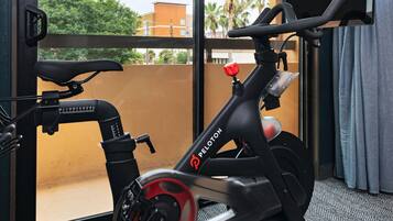 A stationary bike with a red handlebar and a black seat, positioned in a room with a large window overlooking a building and palm trees.