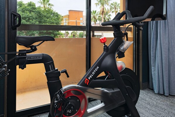 Una bicicleta estática con manillar rojo y asiento negro, ubicada en una habitación con un ventanal grande que da a un edificio y palmeras.