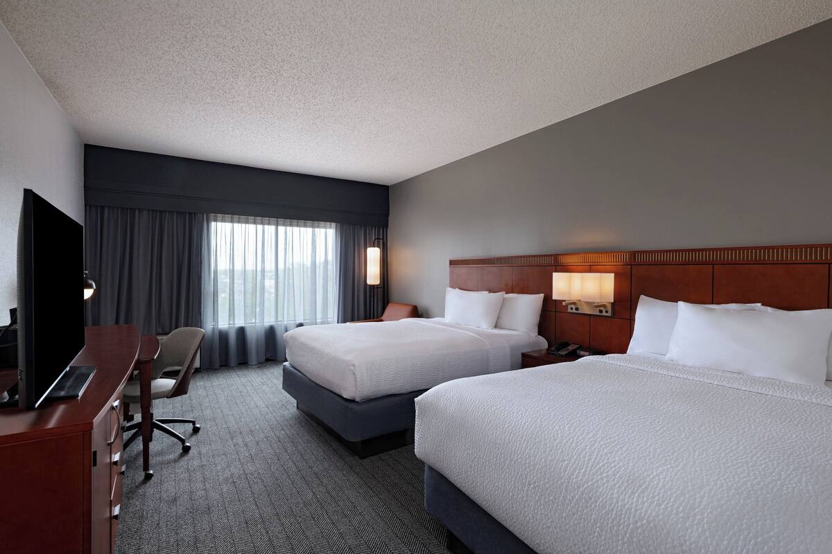 A hotel room with two beds, a desk, a chair, a TV, and a large window with curtains.