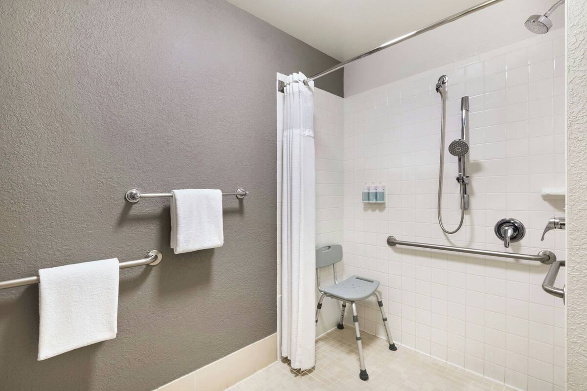 A bathroom with a shower, grab bars, a wheelchair-accessible bench, and a towel rack.