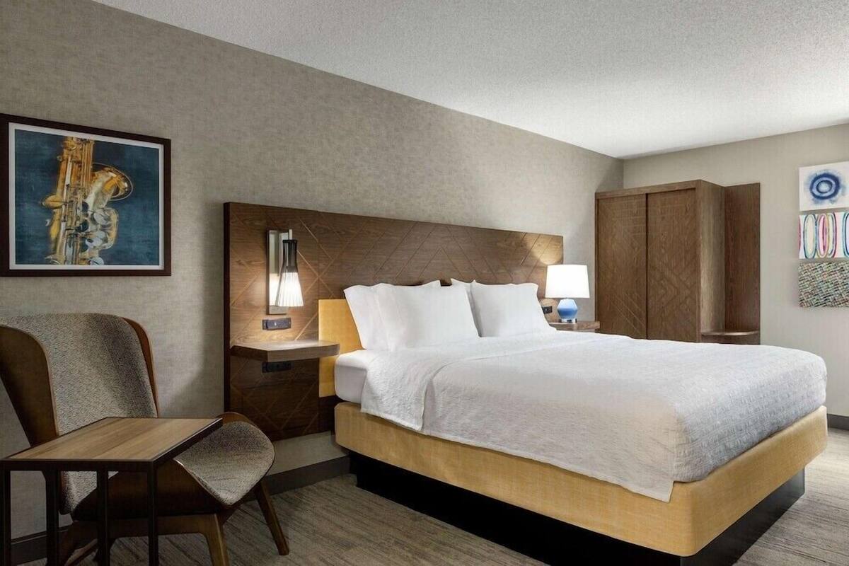 A hotel room with a bed, a chair, a nightstand with a lamp, a wooden headboard, and a painting on the wall.