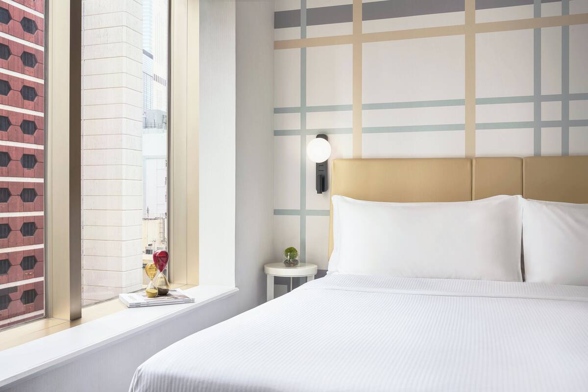 A hotel room with a large bed, a bedside table, a wall clock, and a window with a view of a building.