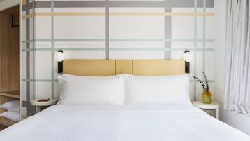 A neatly made bed with white linens, a headboard with a light wood finish, and a small bedside table with a lamp and a vase with a plant.