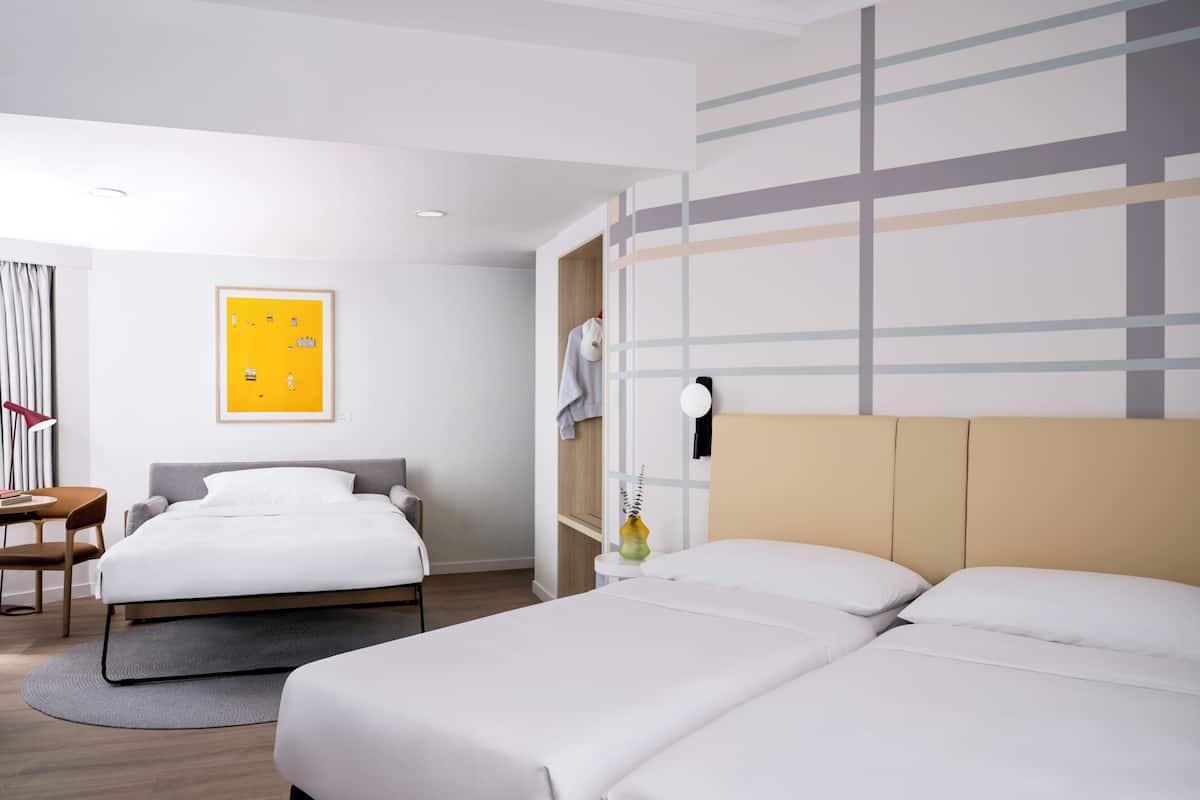 A hotel room with two beds, a desk, a chair, a yellow artwork, and a wall with a geometric pattern.