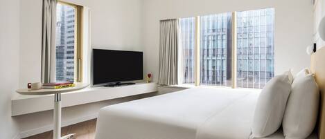Habitación de hotel con una cama grande, un televisor sobre un mueble, una mesita con libros y vistas a edificios a través de las ventanas.