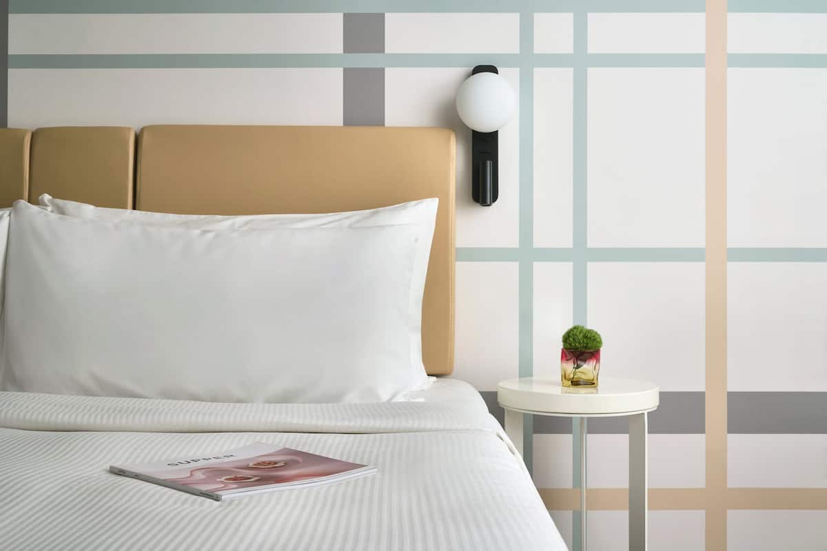 A neatly made bed with a magazine, a wall-mounted lamp, and a small table with a plant.