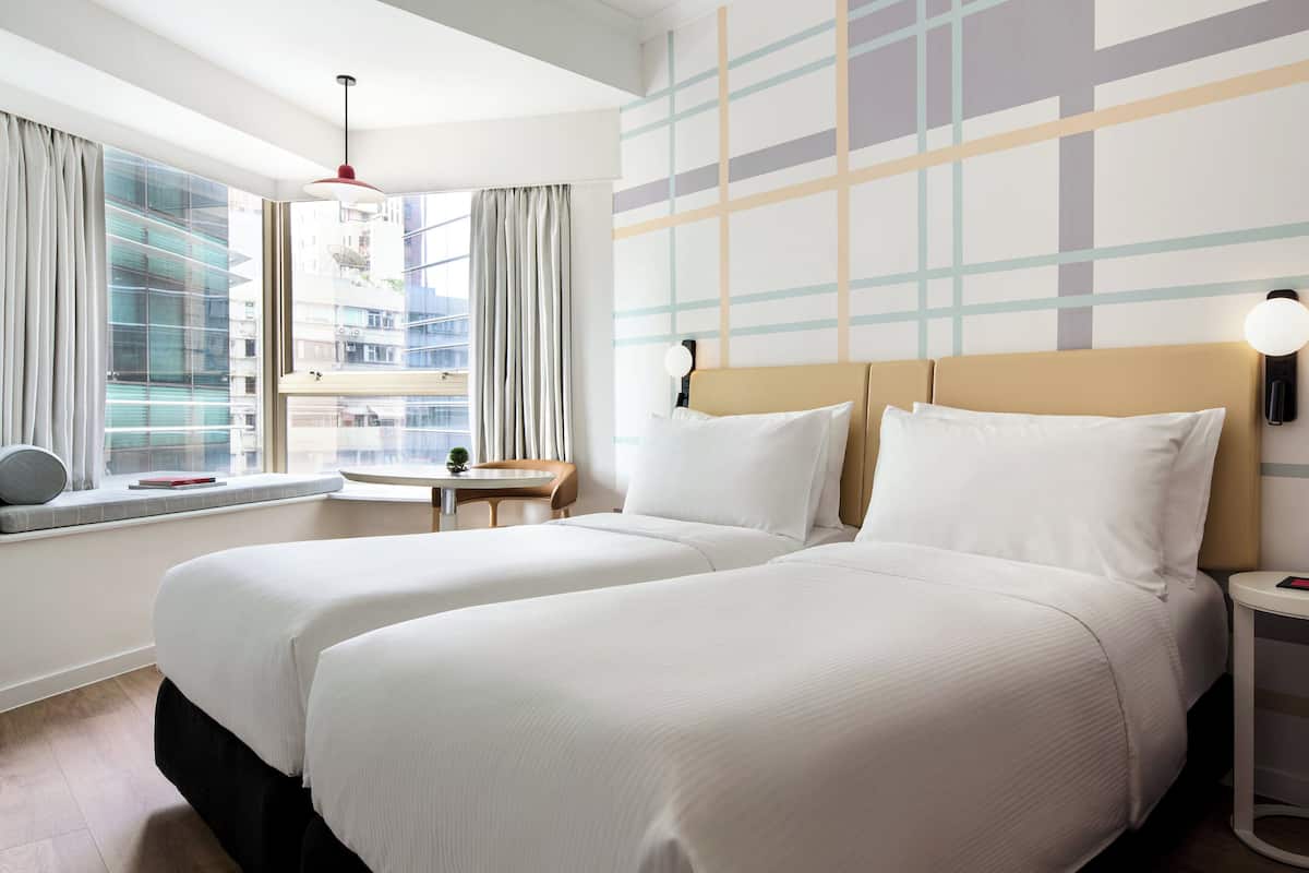 A hotel room with two beds, a window with curtains, a small table, and a wall with a geometric pattern.