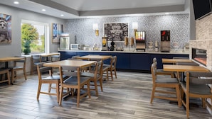 Un café moderno con mesas y sillas de madera, una zona de cocina y una pared de ladrillo con un cartel que dice 'Good Morning'.