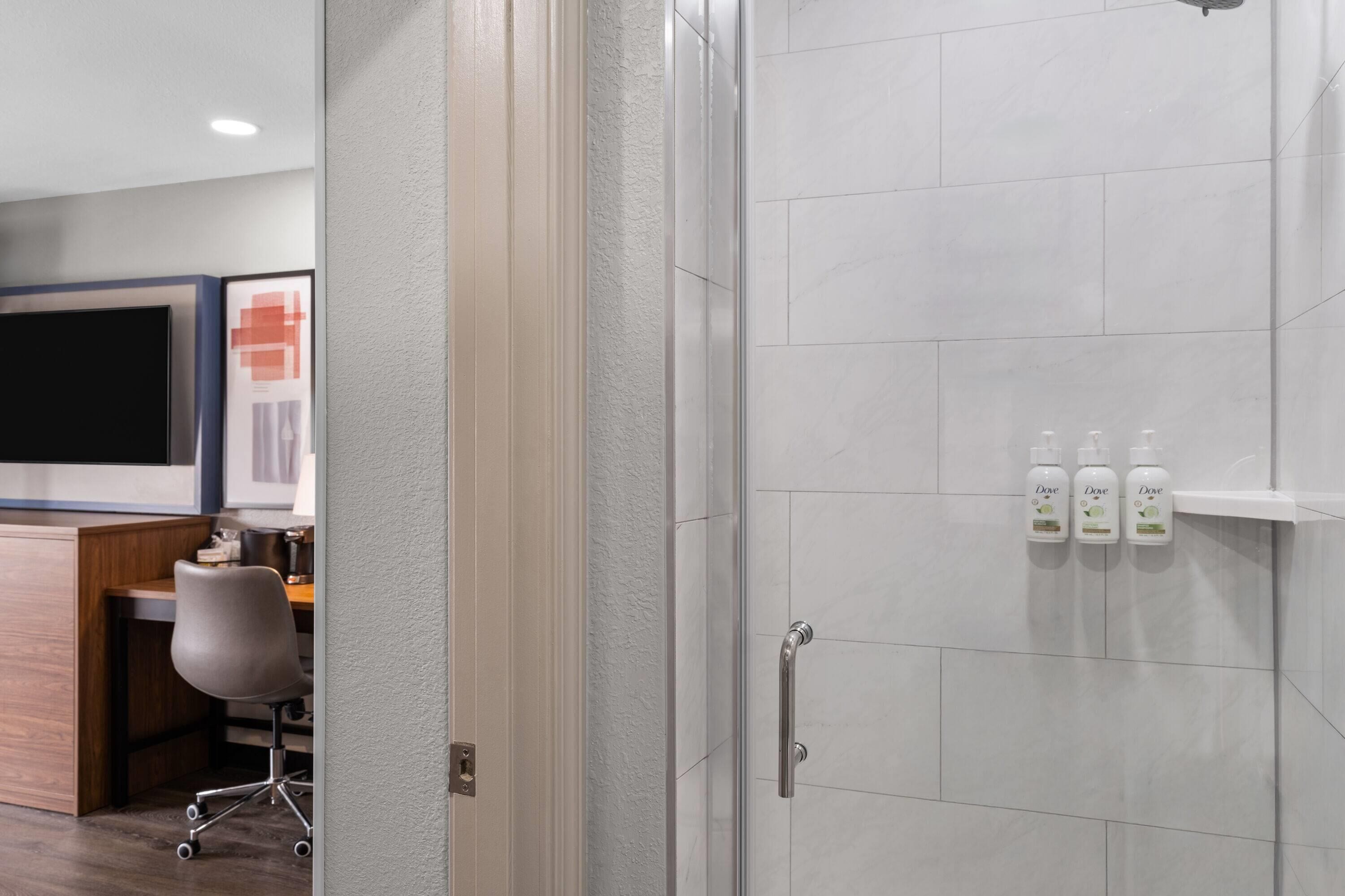 A modern bathroom with white tiles, a shower area, and a shelf with three bottles of Dove body wash.