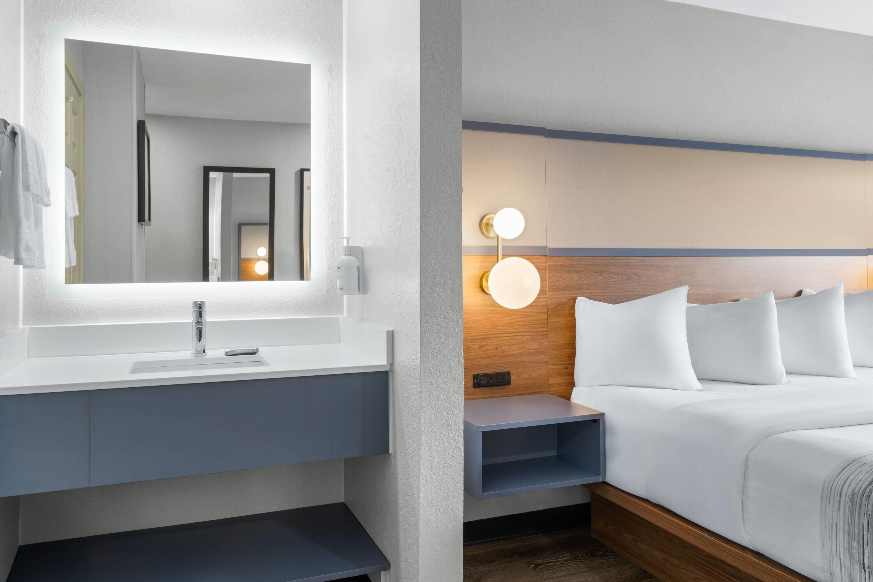 A modern hotel room with a white sink, mirror, bedside table, and a bed with white bedding.