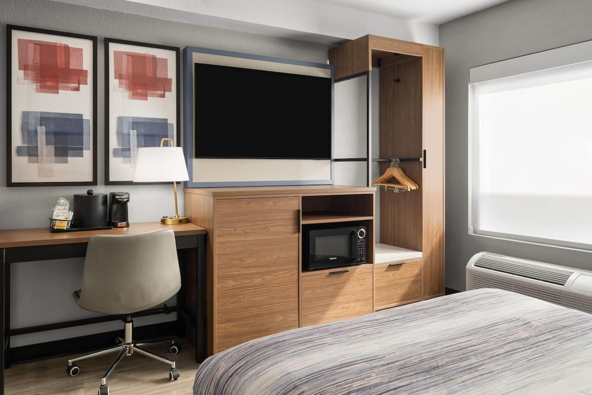 Habitación de hotel con un escritorio de madera, una silla moderna, una cama con ropa de cama a rayas, un televisor de pantalla plana, un microondas, una cafetera y una ventana con persianas.