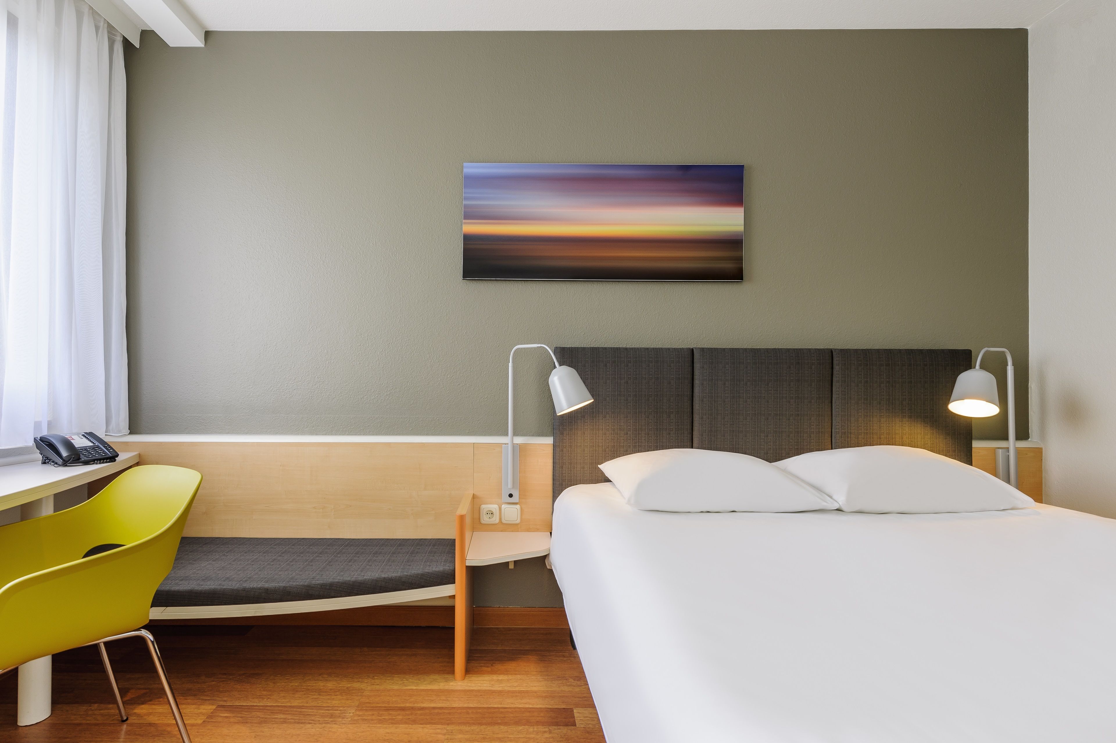 A hotel room with a bed, bedside lamps, a desk with a chair, a small table, and a wall-mounted phone.