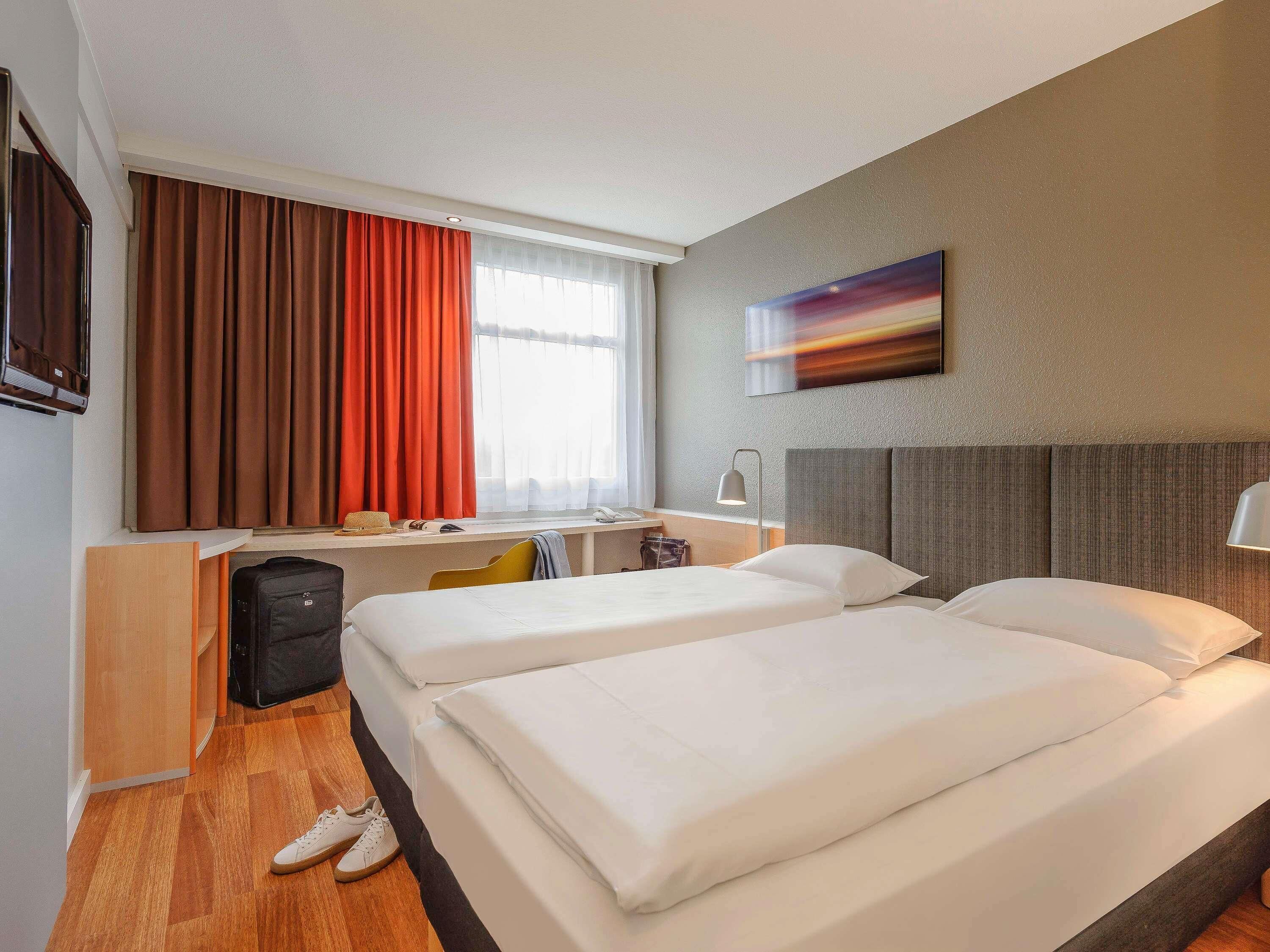 A hotel room with two beds, a desk, a chair, a TV, and a painting on the wall.