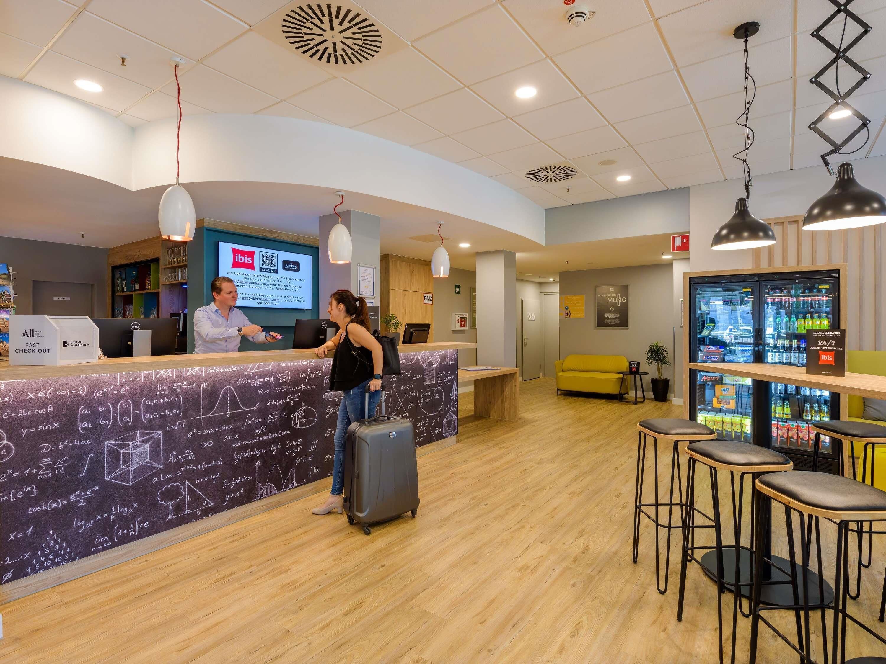 Ein Hotel-Empfangsbereich mit Theke, digitalem Bildschirm, einer Sitzecke mit gelber Couch und einem Getränkekühlschrank.