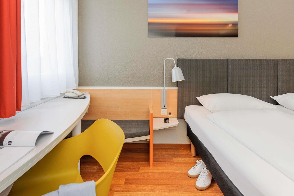 Una habitación de hotel con una cama, un escritorio, una silla, una lámpara y un cuadro en la pared.