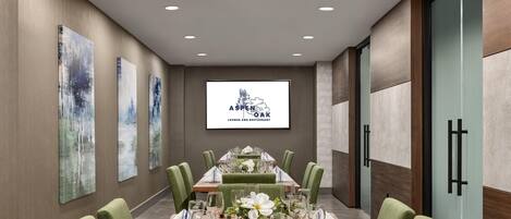 Un salón de conferencias con una larga mesa, sillas tapizadas de verde y una pantalla proyectora mostrando 'Aspen Oak Lounge and Restaurant'.
