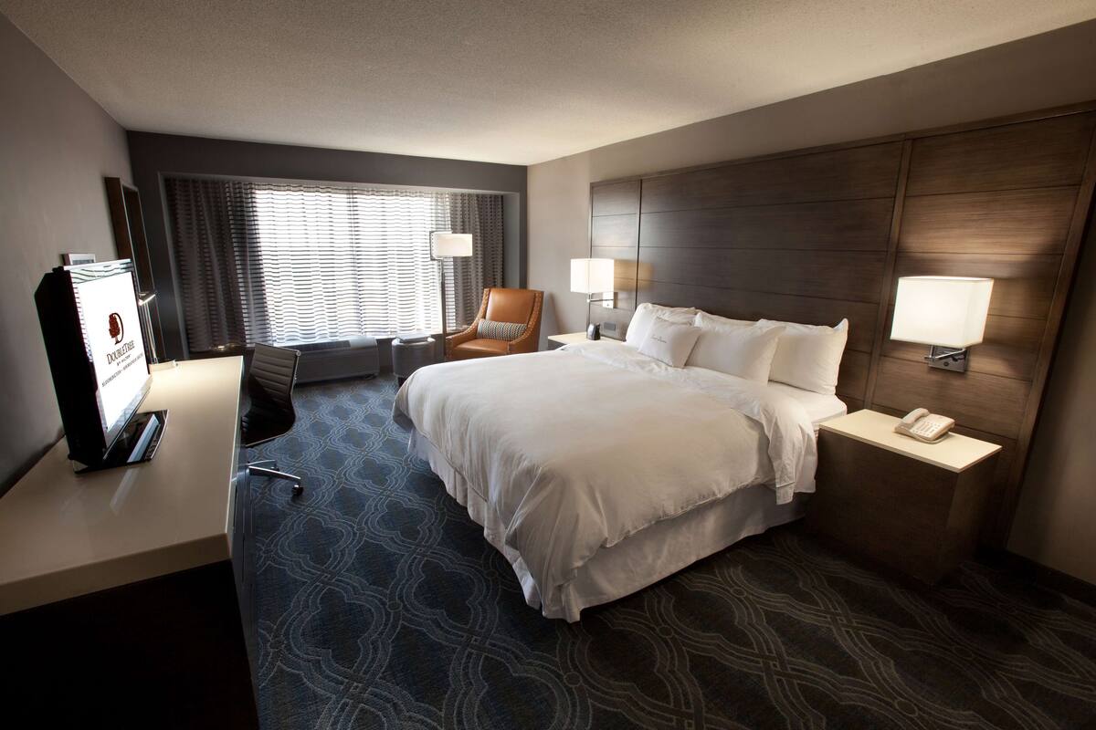 Een hotelkamer met een groot bed, twee bedlampen, een bureau met telefoon, een stoel en een raam met gordijnen.