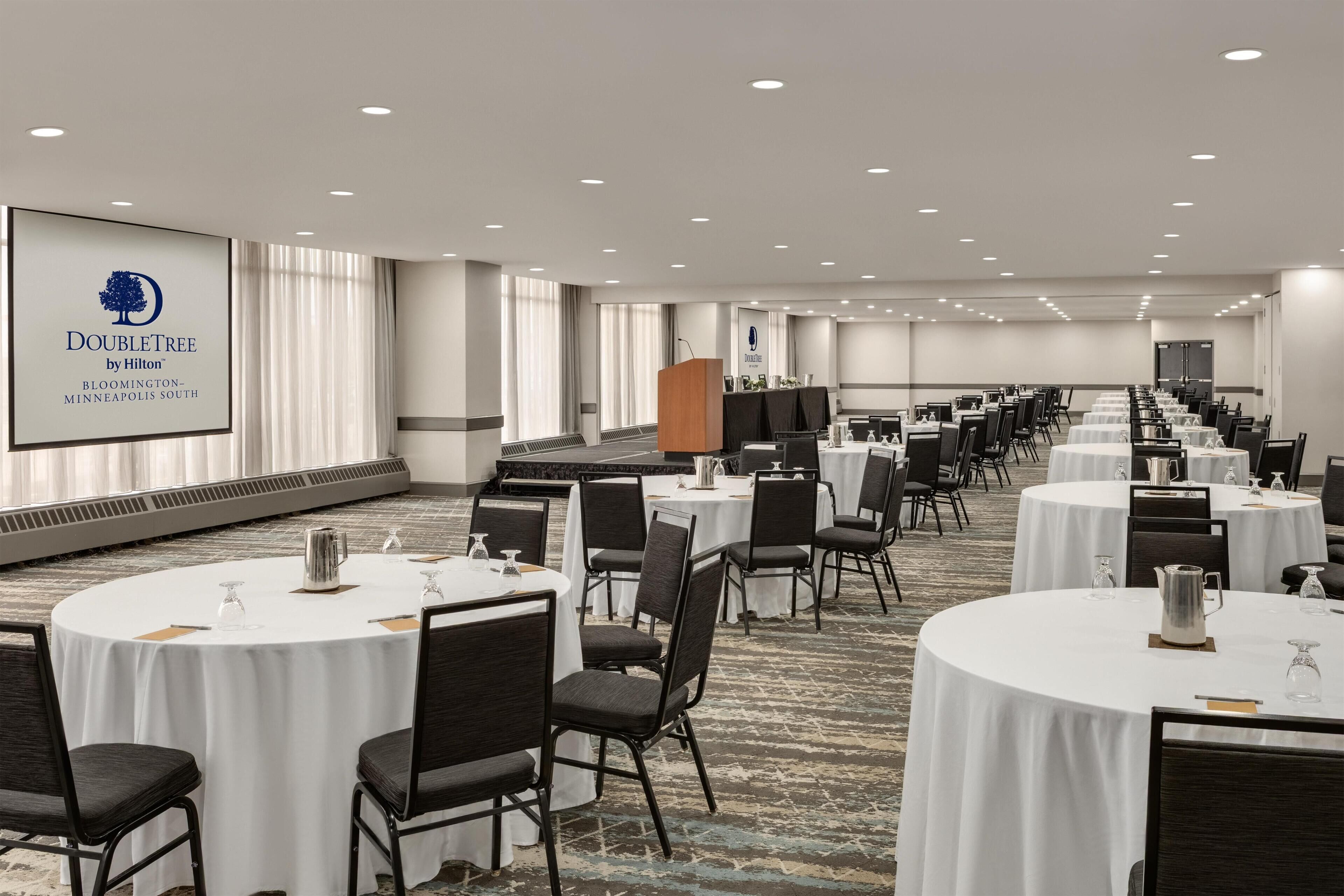A hotel conference room with multiple round tables, chairs, and a large screen displaying the hotel's name.
