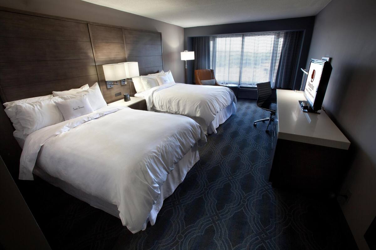 Hotelkamer met twee bedden, een bureau, een stoel, een televisie en een raam met jaloezieën.