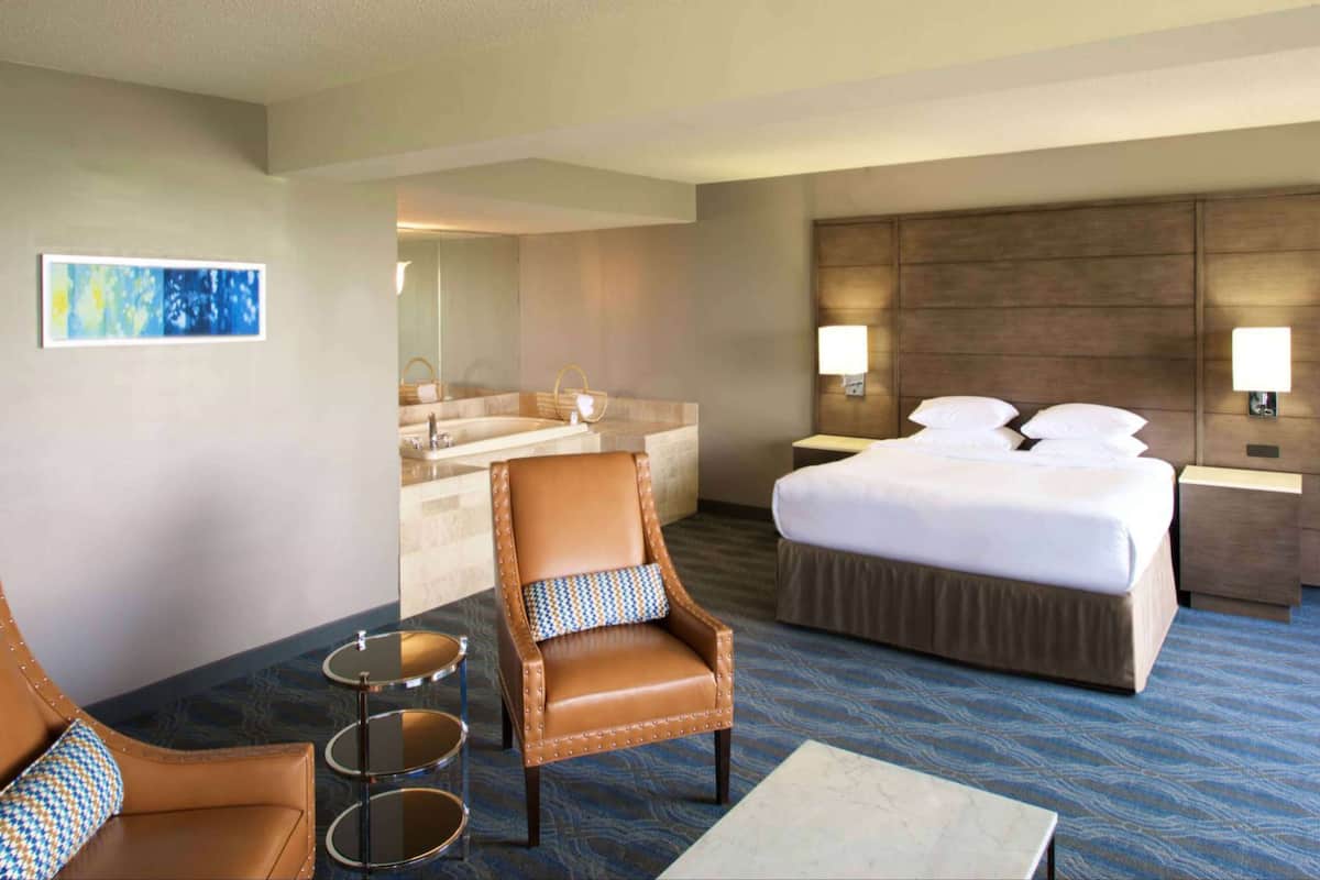 Een hotelkamer met een bed, een stoel, een klein tafeltje, een badkamer met een bad en een wandlamp.