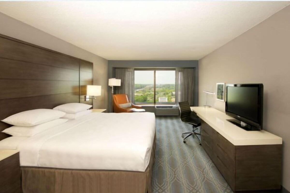 Een hotelkamer met een bed, een bureau met een flatscreen televisie, een stoel en een groot raam met uitzicht op de stad.