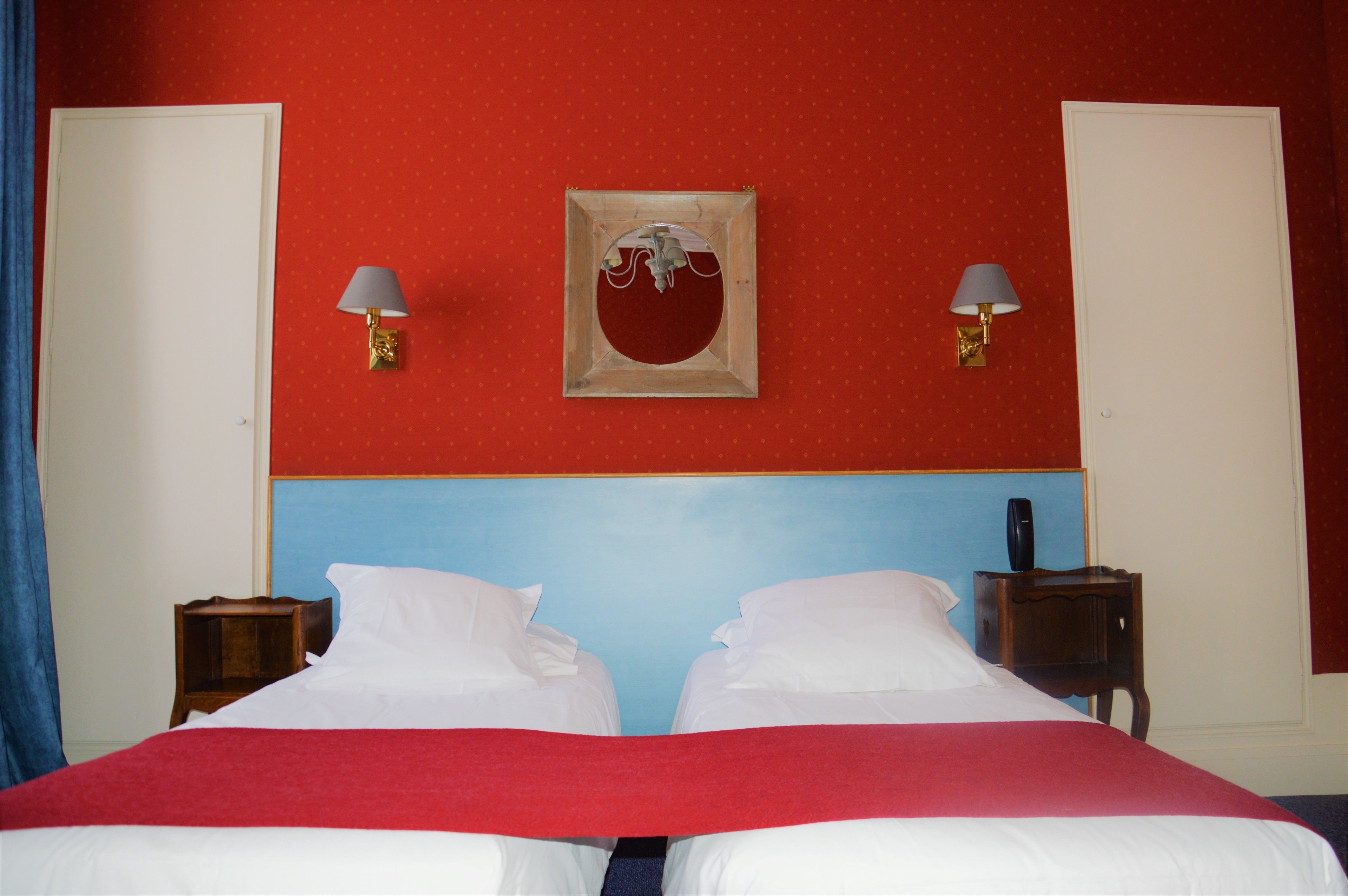 A hotel room with a red and blue color scheme, two beds, a framed artwork on the wall, and two bedside tables with lamps.