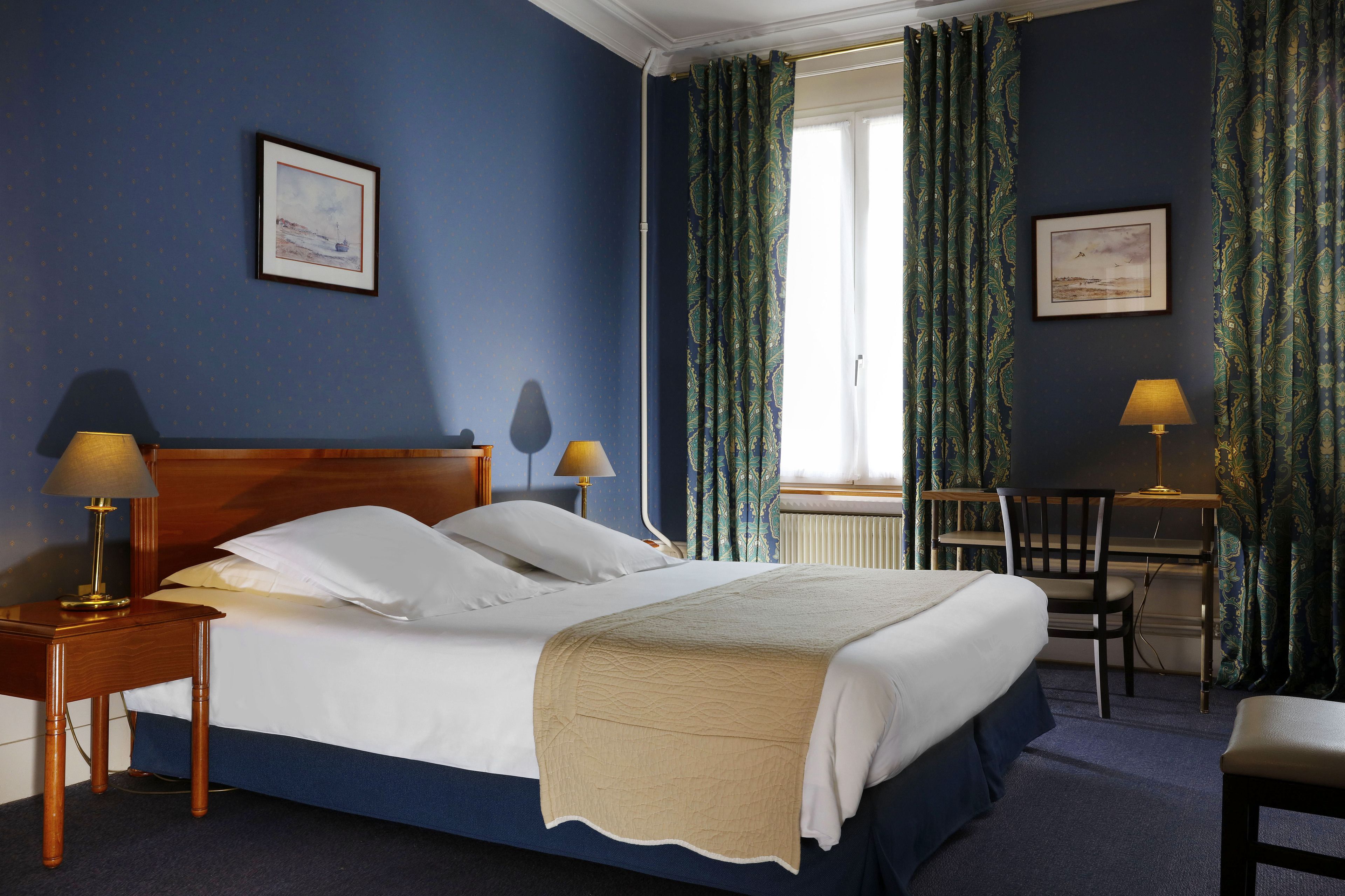 A neatly made bed with white linens, a wooden headboard, and a beige throw blanket. There is a wooden nightstand with a lamp on it to the left of the bed. A desk with a chair and a lamp is positioned to the right of the bed.