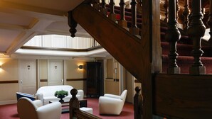 Eine Hotellobby mit einer imposanten Treppe, einer Sitzecke mit zwei Sesseln und einem Sofa sowie einem großen Holzschrank.