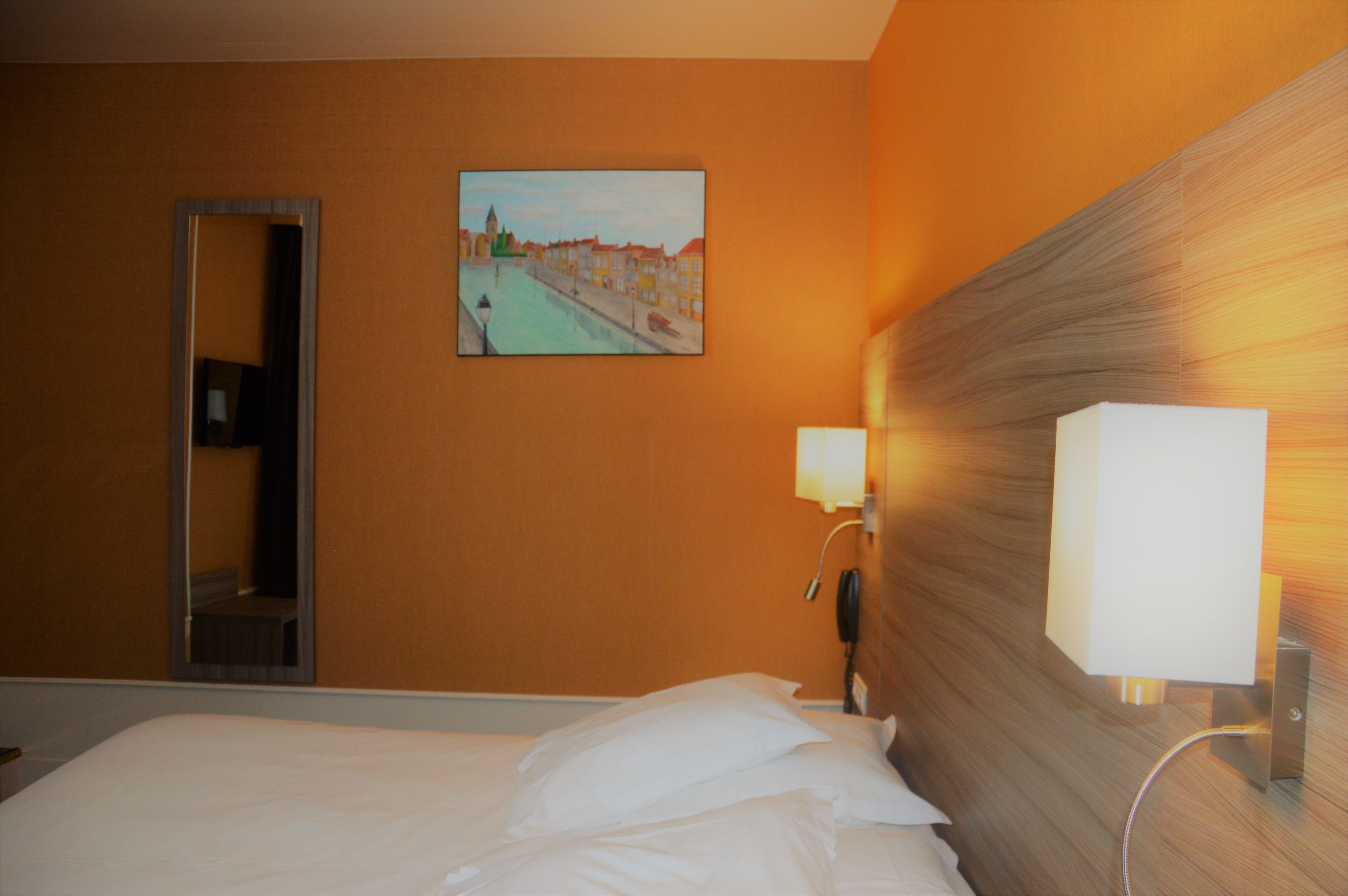 Una habitación de hotel con cama, un cuadro de un paisaje urbano, un espejo y una lámpara de noche.