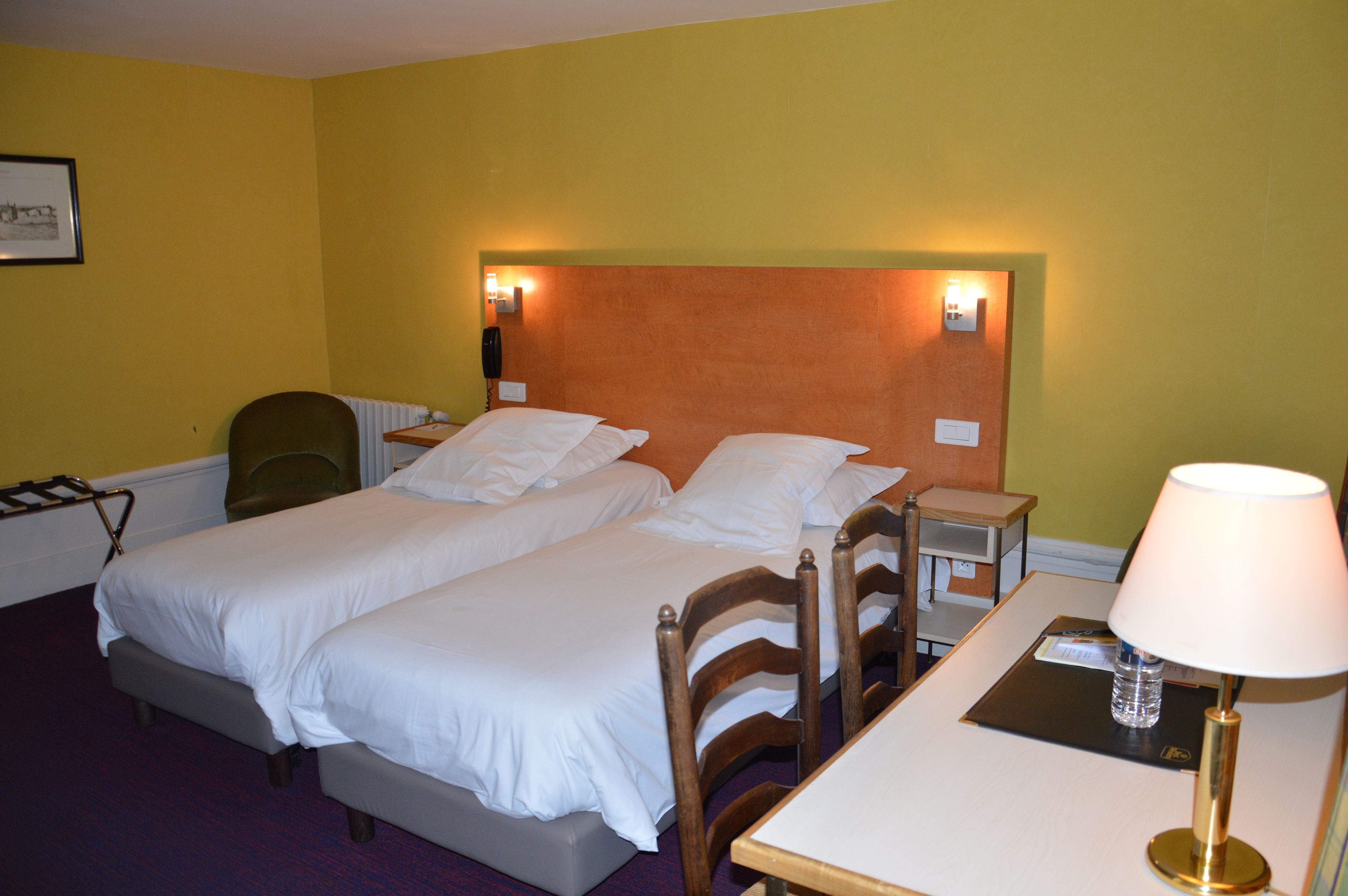 A hotel room with two single beds, a table with a lamp, a chair, and a small desk with a telephone.