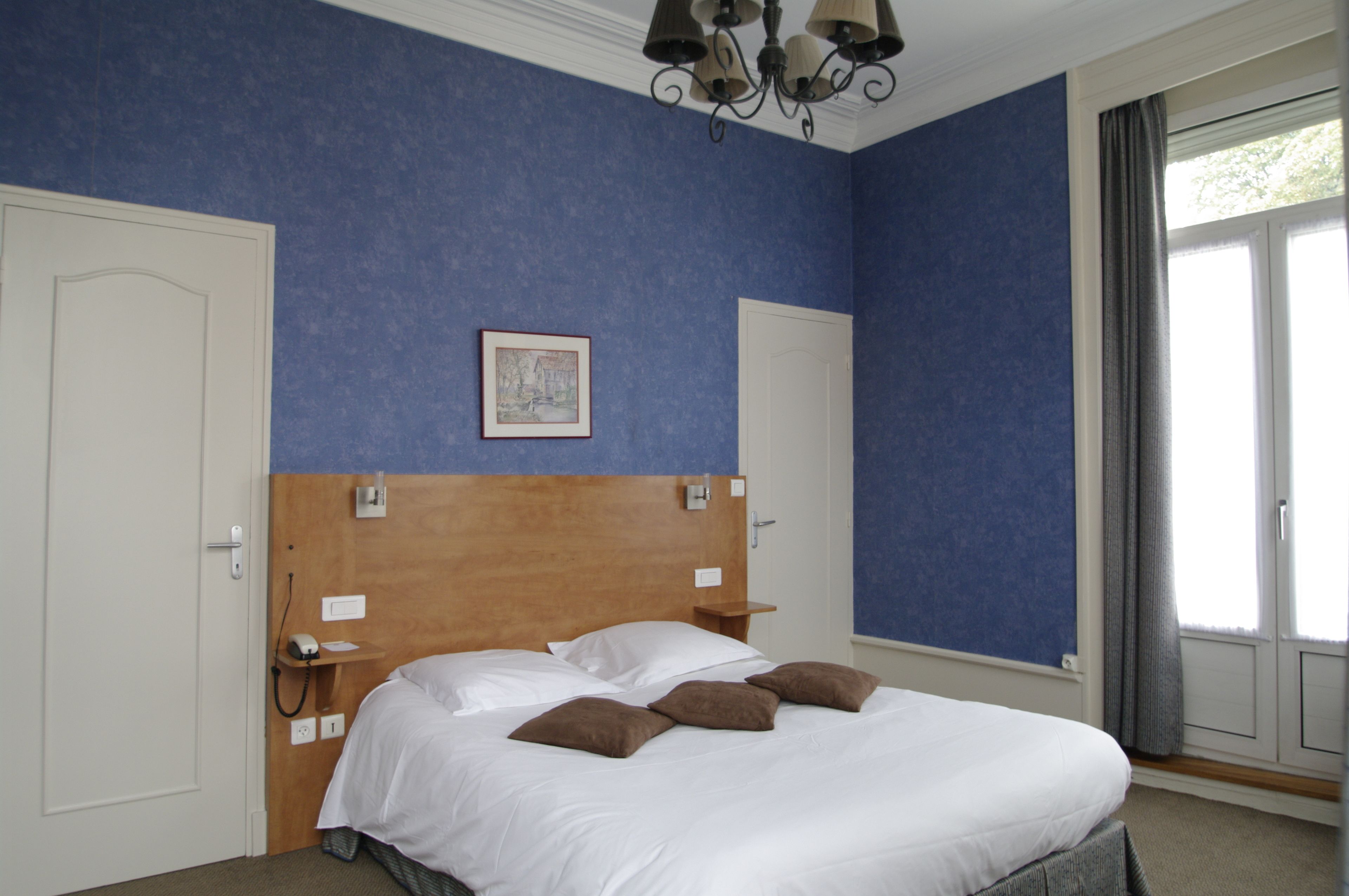 Una habitación de hotel con una cama grande, dos puertas, una araña y una ventana con cortinas.