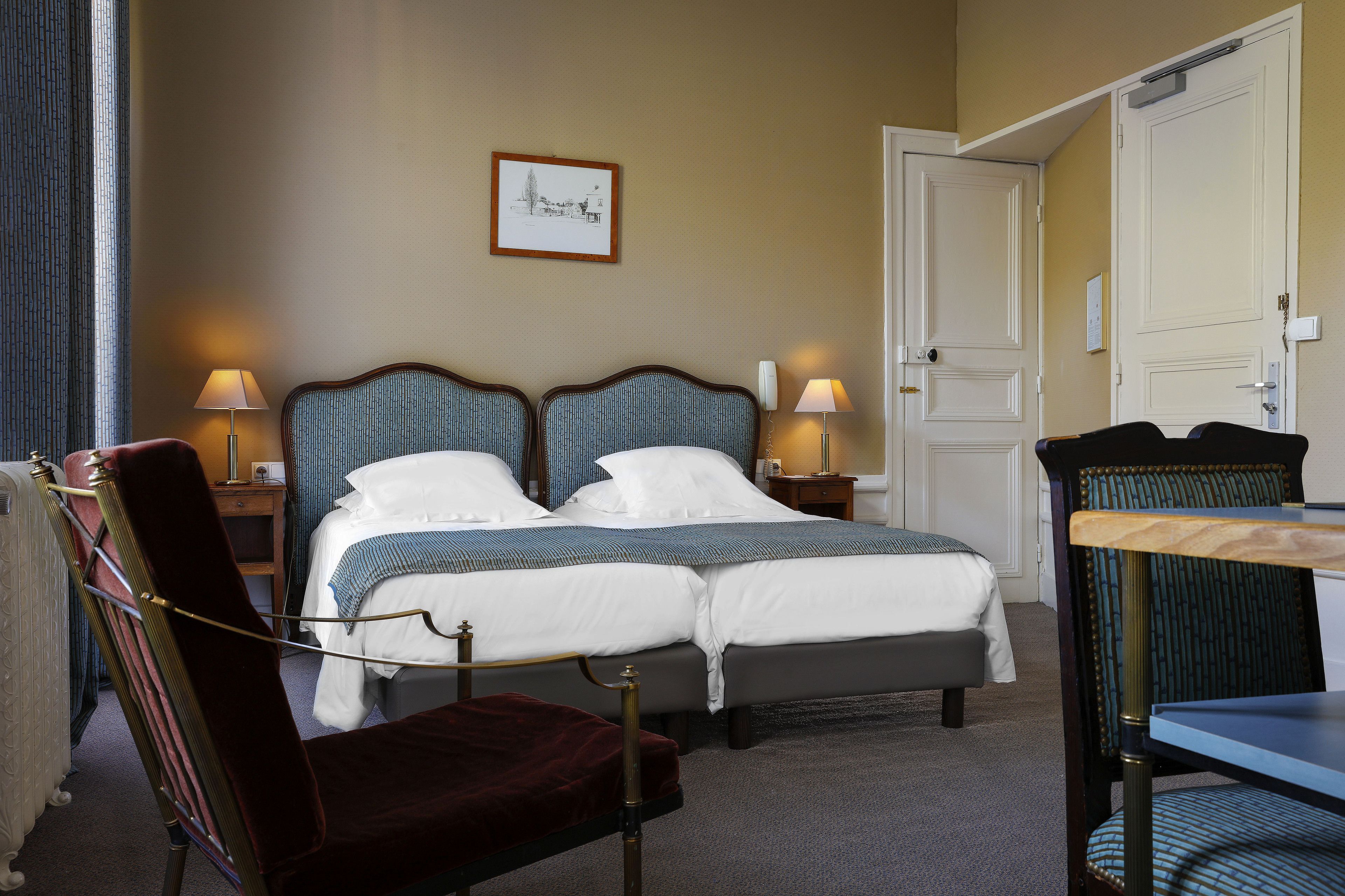 A hotel room with a double bed, two bedside tables with lamps, a chair, a table, and a door.
