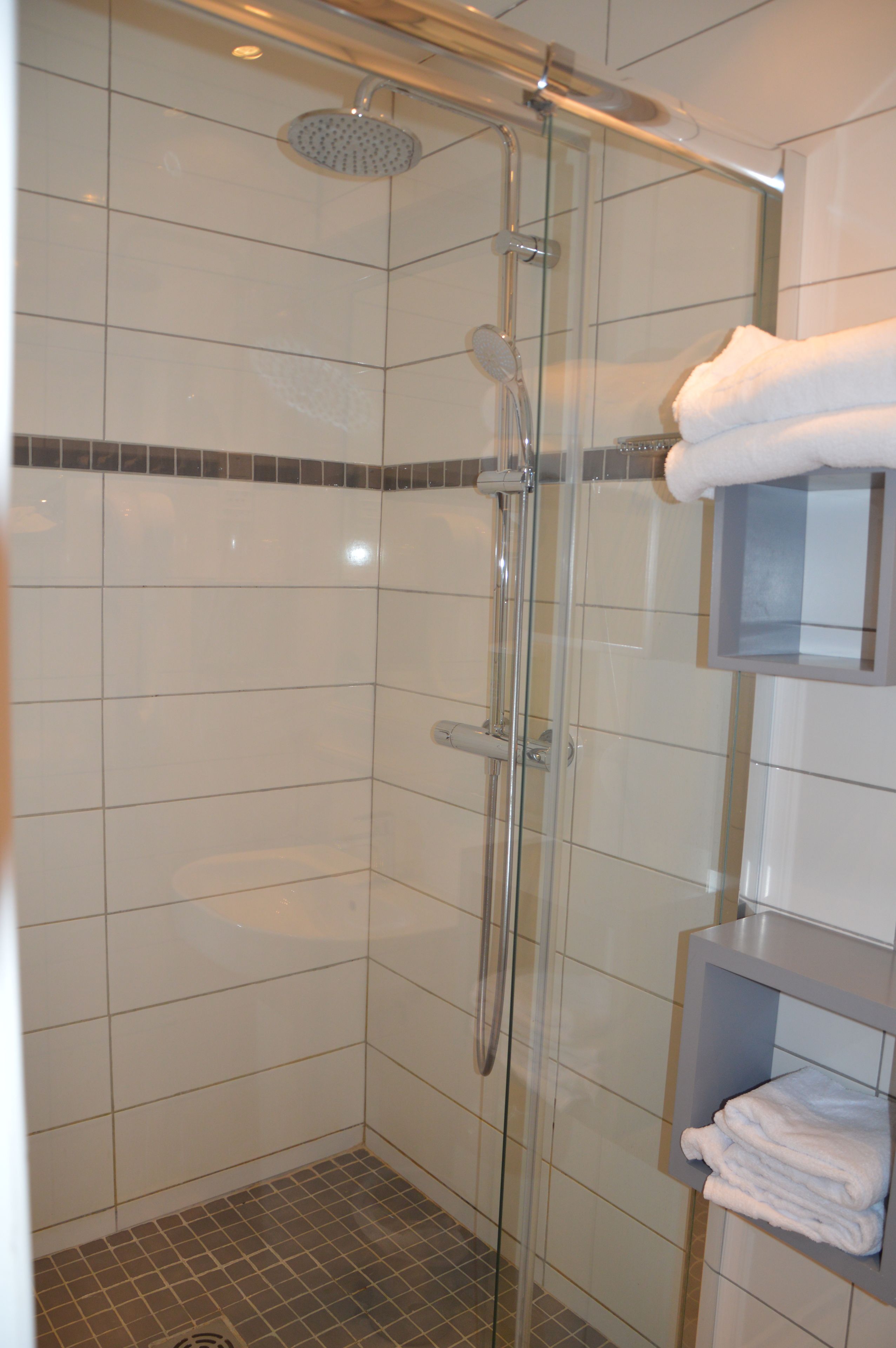 Eine Duschkabine mit Glaswänden, ein Handbrausekopf, ein an der Wand befestigter Handtuchhalter mit gefalteten Handtüchern und ein gefliester Boden.